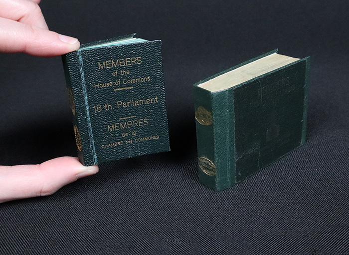 Deux petits livres à reliure vert foncé sont posés debout. Le plus petit est tenu entre le pouce et l’index pour donner un ordre de grandeur.