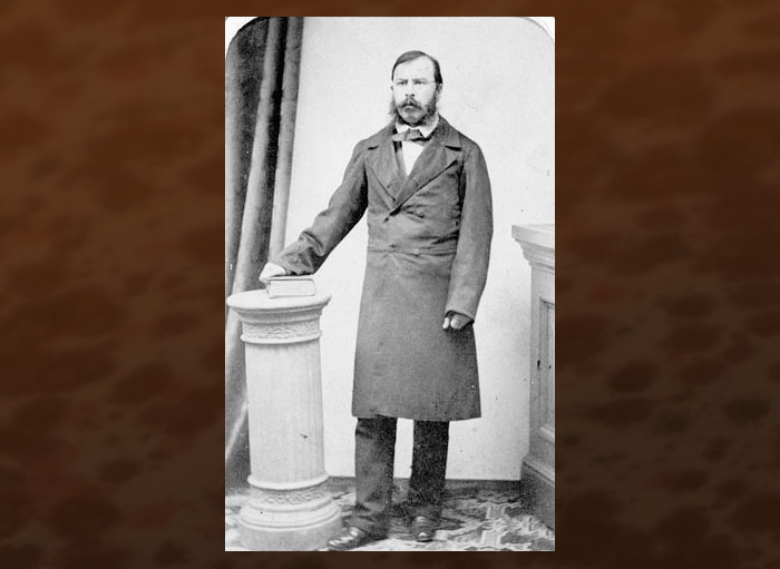 Black and white photograph of Joseph Cauchon in 1862, wearing a long jacket, standing beside a plinth with a book 